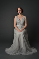 Fototapeta premium full length portrait of brunette girl wearing long silver ball gown. seated pose on grey studio background.