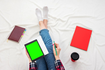Girl in blue jeans and plaid shirt, woman holding a tablet pc and notebook, women's feet, lying on white crumpled blanket. Women's hands. Background with copy space, for advertisement. Top view