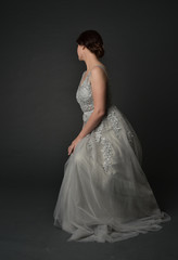 Fototapeta premium full length portrait of brunette girl wearing long silver ball gown. seated pose on grey studio background.