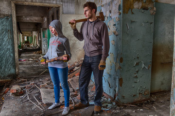 A pair of hooligans: a girl in hood with a bat and a guy with a sledgehammer, standing in the middle of the ruins of an old building. Maybe street band or fighters