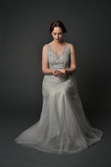 Fototapeta premium full length portrait of brunette girl wearing long silver ball gown. seated pose on grey studio background.