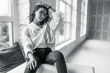 young cute sexy charming girl in a white man's shirt and dark jeans sitting by the window on a...