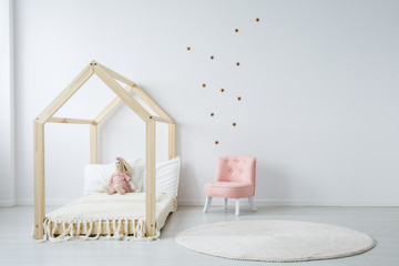 Pastel pink armchair next to wooden house shape bed with toy and blanket, copy space and golden...