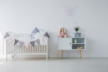 White wooden crib with pillows next to wooden cabinet with toy and green pant in grey, copy space...