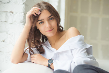 young cute sexy charming girl in a white man's shirt and dark jeans sitting by the window on a...