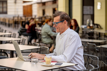 Happy attractive stylish mature man working on laptop while having coffee in outside coffee shop