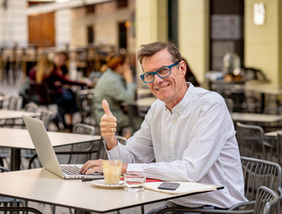 Fototapeta premium Happy attractive stylish mature man working on laptop showing thumb up in outside coffee shop