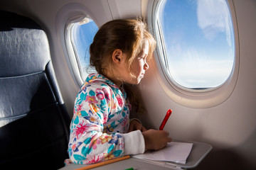 Fototapeta na wymiar Child drawing picture with crayons in airplane. Little girl occupied while flying in aircraft. Travel with family and kids. Blue sky and sun outside the window
