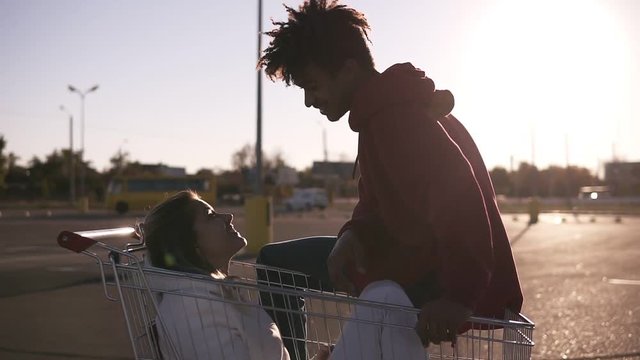 Afroamerican young boy with trendy haircut sitting on a shopping cort on the ouside parking zone and looking to his girlfriend. Caucasian girlfriend sitting inside the cort and talking to her