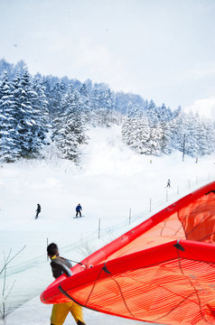 A superimposed or double exposures of a man carries his red windsurf in a ski resort with people having fun skiing down the mountain. This conceptual image showing a surprise holiday; winter & summer.
