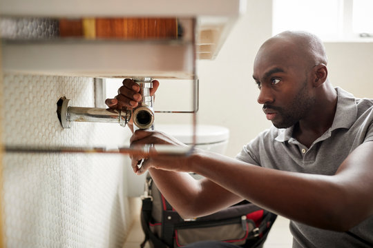 Young black male plumber sitting on the floor fixing a bathroom sink, close up