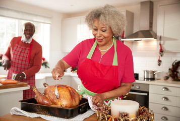 Senior black woman basting a roast turkey in preparation for Christmas dinner, her husband chopping...