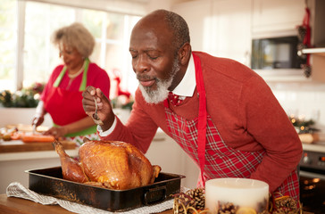 Senior black man basting roast turkey in preparation for Christmas dinner, his wife chopping...