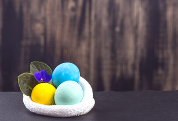 Colored chicken eggs lie in a knitted bag on a wooden background, flowers of violets, the concept of the background for the design for Easter