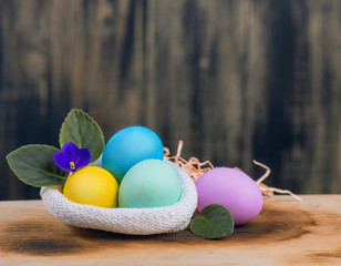 Colored chicken eggs lie in a knitted bag on a wooden background, flowers of violets, the concept of the background for the design for Easter