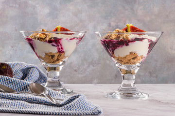 Figs pudding parfait with yogurt, blueberry jam, figs, hazelnut, and cookies in glass on white marble background.