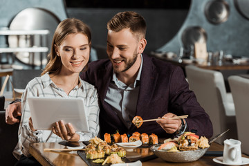 Attractive couple using digital tablet while eating sushi in restaurant