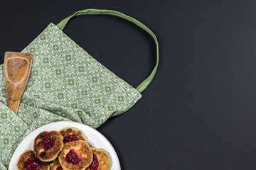 Green apron, wooden spatula and plate with cotton cheese pancakes on a dark background.