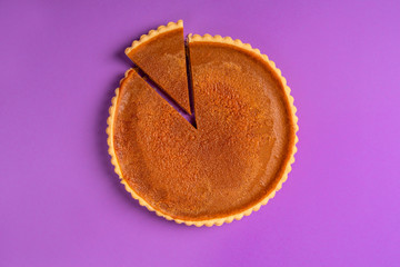Whole pumpkin pie with a cut slice. Above view. Purple background.