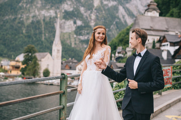 Fototapeta premium A beautiful wedding couple walks on a background old cathedral, lake and mountains in a fairy Austrian town, Hallstatt.