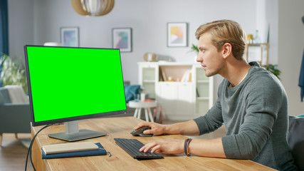 Handsome Young Man Works on a Green Mock-up Screen Personal Computer while Sitting at His Desk in...