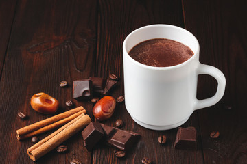 Hot chocolate drink in a white cup, chocolate cubes, cinnamon sticks and coffee bean on the dark wooden background