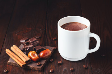 Hot chocolate drink in a white cup, chocolate cubes, cinnamon sticks and coffee bean on the dark wooden background