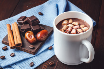 Hot chocolate drink with marshmallows in a white cup, chocolate cubes, cinnamon sticks and coffee bean on the dark wooden background