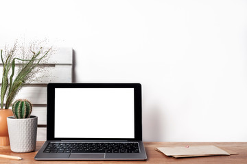 Hipster wooden desk with an open laptop mockup, cactus in a gray pot, boards on the wall, a brown vase with green grass and a white empty wall copy space