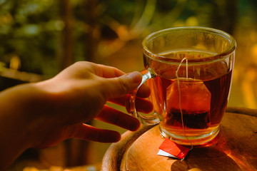 A hand lifting a glass cup of black tea on a table with autumn trees in the background.