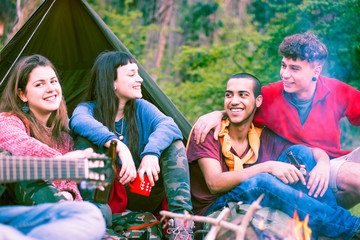 Group of multiracial friends sitting at campfire playing music -  Happy multicultural teenagers having fun around fire drinking together on vacation - Concept of  friendship and outdoor activity 