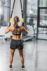 back view of muscular sportswoman working out with suspension straps in gym