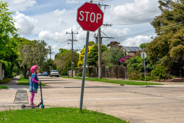 Young girl in pink outfit and pink helmet riding a kick-scooter, waiting at crossing with stop sign