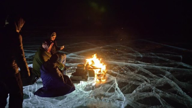 Four travelers by fire right on ice at night. Campground on ice. Tent stands next to fire. Lake Baikal. Nearby there is car. People are warming around campfire and are dressed in sleeping bags