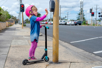 Young girl in pink outfit and pink helmet riding a kick-scooter pressing button at pedestriam crossing