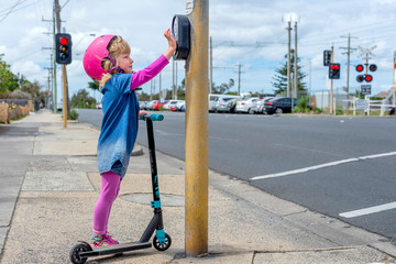 Young girl in pink outfit and pink helmet riding a kick-scooter pressing button at pedestriam crossing