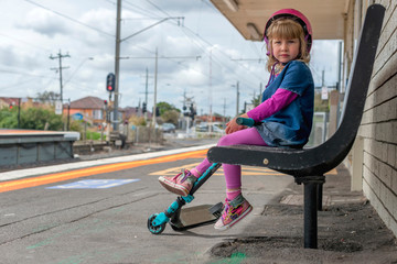 Young girl in pink outfit and pink helmet riding a kick-scooter waiting on a platform at train station