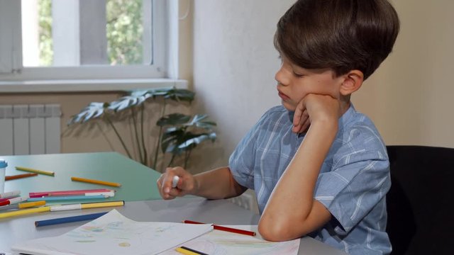 Young schoolboy looking bored while coloring his sketchbook. Cute little boy sitting alone at the classroom, drawing and coloring. Boy working on art project looking tired. Kids education concept.