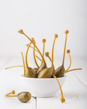 A bunch of marinated (pickled) capers in a bowl on white wooden background.  Edible fruits of Capparis. Caper berries are used as a garnish. Capers with selective focus, macro, close up. Copy space.
