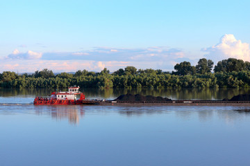 Tugboat pulling heavy loaded barge of black coal along the green trees on the shore of Danube river