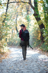 Female hunter walks on the road in the forest/Young woman with gun goes on a stone road in the forest. View from the back.