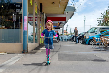 Young girl in pink outfit and pink helmet riding a kick-scooter on sidewalk