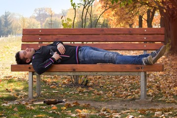A drunk young man sleeping on a bench in the day.A young man holding a bottle of alcohol in her...