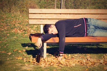 A drunk young man sleeping on a bench in the day.A young man holding a bottle of alcohol in her...
