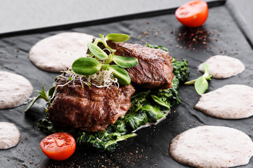 Medallions of veal on a spinach pillow with sprouts of grain, cream sauce and tomatoes on a black plate on a dark background. Beautiful serving of food. Flat lay. Space