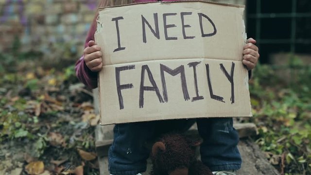Homeless child outdoors. Holding cardboard with sign i need family on it.