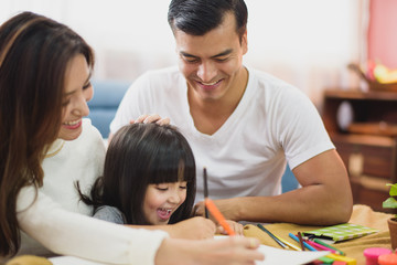 Fototapeta premium Family time Daughter laughing enjoying writing painting with her father and mother on the table in front of sofa at home.