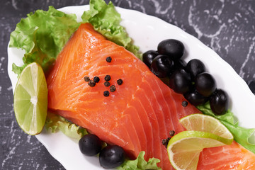 A piece of salmon on a white plate with olives and lemon, on a gray background. Selective focus