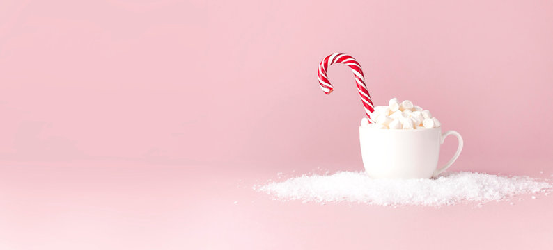Christmas New Year drink, white mug with marshmallows and Candy Cane in the snow on pink background Flat Lay copy space. Winter traditional drink food. Festive decor, celebration Xmas holiday 2019