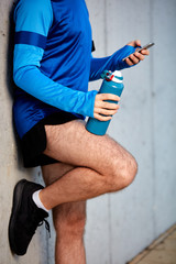 Close up of male runner leaning on wall and using smart phone. In other hand bottle with water.